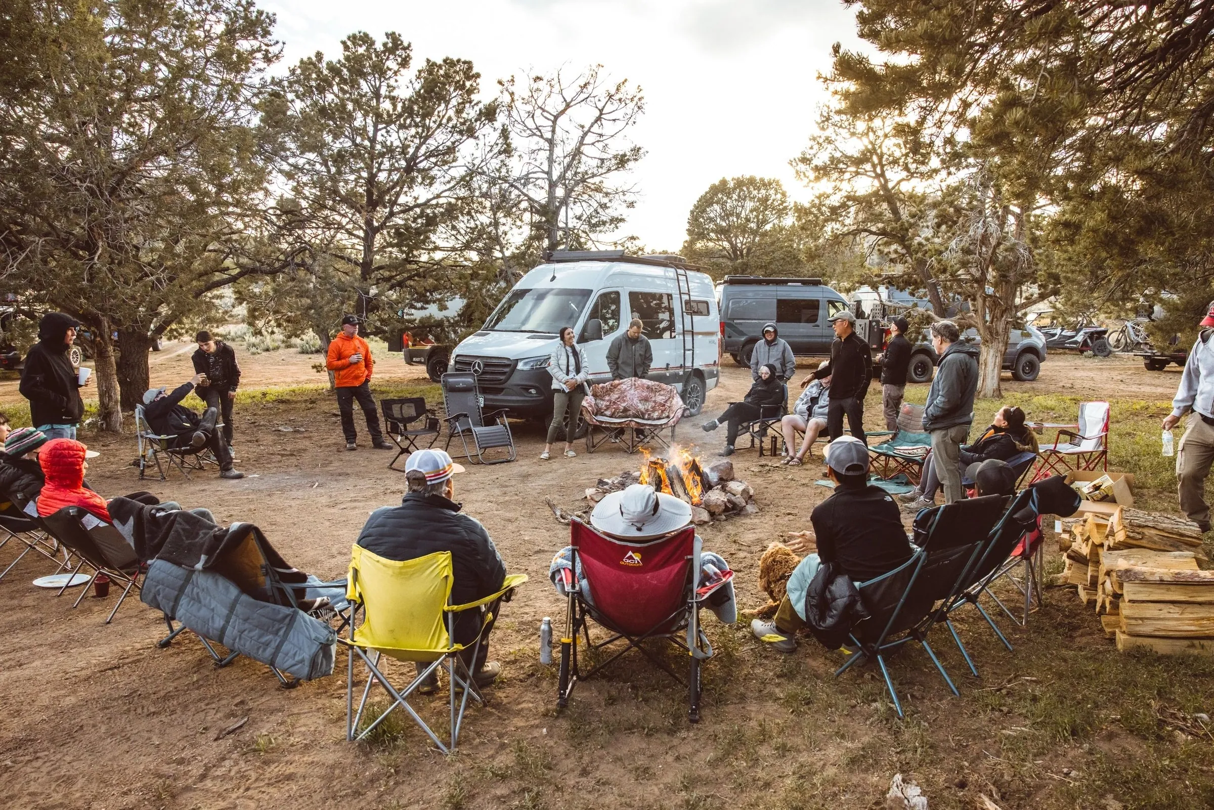 Guests relaxing in camp chairs with camper vans parked among pinyon trees