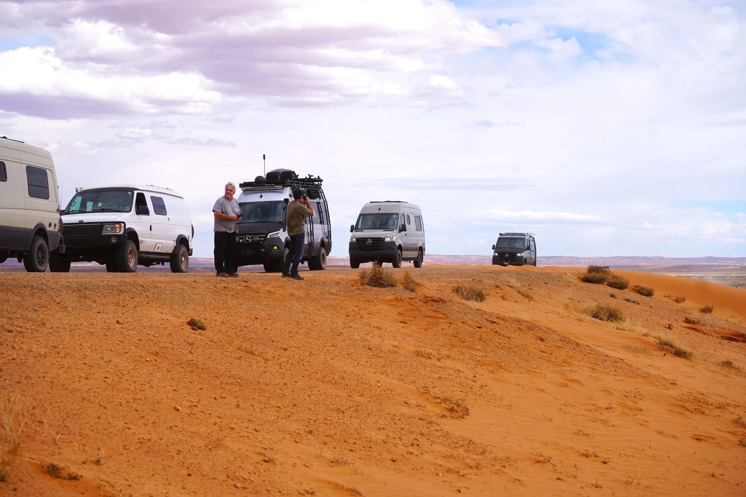 Camper vans and trucks gathered on red sand under a cloudy sky