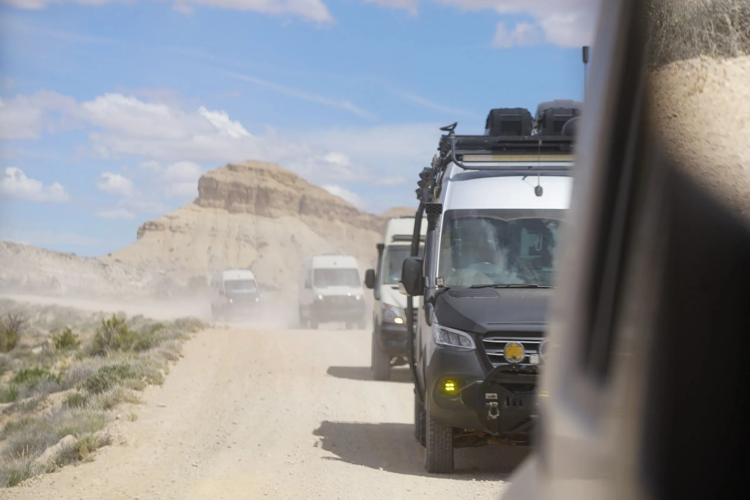 Camper vans in a dusty convoy on a gravel road past pale cliffs